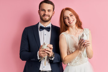 Portrait of caucasian man and woman holding two white pigeons isolated over pink background. Redhaired woman in dress and bearded man in tux looking at camera