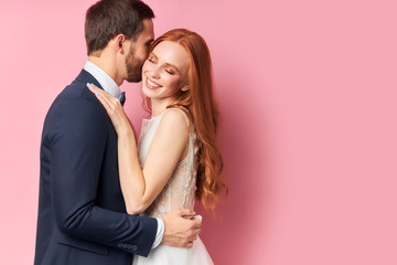 Caucasian man in suit and woman in white wedding dress with long hair lovely hug each other and smile, after wedding ceremony posing isolated over pink background.