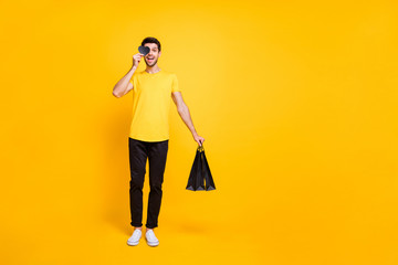 Full length photo of handsome guy holding boutique bags little paper heart postcard making surprise girlfriend wear casual t-shirt pants isolated yellow color background