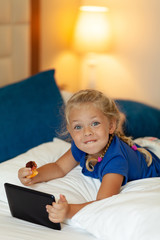 Little pretty girl holding tablet in her hands eats fruit and lies on bed (sofa) at home. Mobile game, entertainment and communication via Internet.