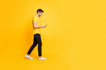 Fototapeta na wymiar Full length body size photo of cheerful attractive man with bristle smiling toothily focused on reading feednews on his phone wearing yellow t-shirt white footwear isolated bright color background