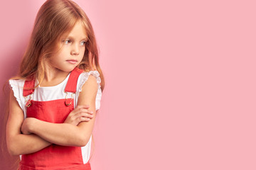 Upset caucasian girl wearing red overalls stand isolated over pink background. Looking side with crossed arms
