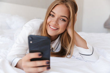 Photo of cheerful woman using cellphone and smiling while lying on bed