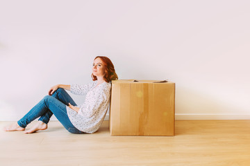 Young attractive girl moving into the new apartment. Sitting on the floor with big carton box in an empty room on a white wall background.