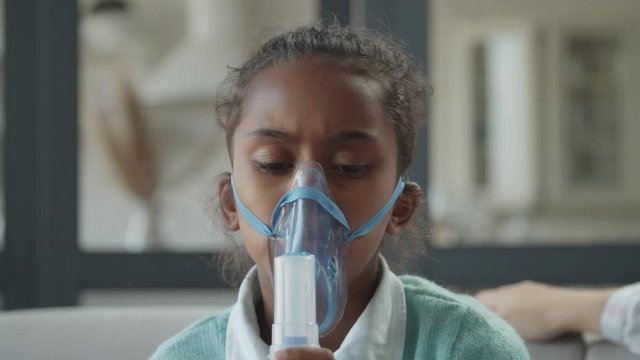 Portrait of beautiful african american elementary age girl doing asthma breathing treatment with nebulizer at home. Sick cute child making inhalation with medical ultrasonic inhaler in domestic room.