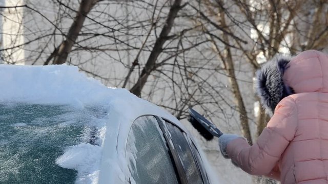 Winter portrait of a young woman cleaning snow from a car. Beauty blonde Model Girl laughs and cheerfully cleans the snow. Beautiful young woman outdoors.