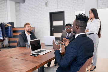 Young business people consisted of caucasian and african gathered to speak. Everyone listening to african speaker man with laptop talking about new business project