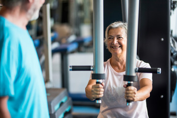 couple of two seniors at the gym training together - woman doing a exercise in a machine and the...