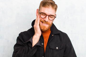 young red head man holding cheek and suffering painful toothache, feeling ill, miserable and...