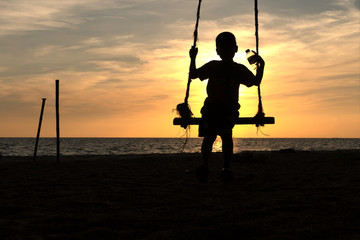 Silhoutte image of young boy having fun on the swing with water bottle in hand, kid playing wooden swing on the beach in sunset.