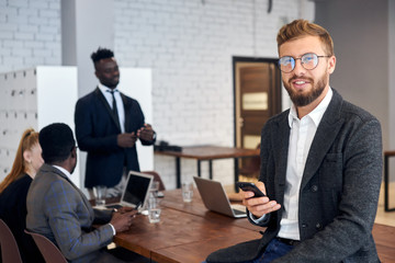 Cheerful young man in tuxedo and eyeglasses using smartphone in workplace while his business colleagues having conversation in background