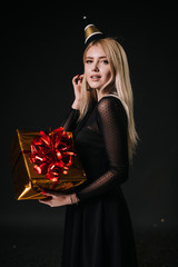 Beautiful young woman in a festive cap holds a holiday box with a gift. Girl blonde is seductively looking at the camera. Shooting in professional studio on isolated black background.