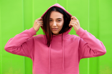 Fototapeta na wymiar Portrait of young teenage girl smiling and looking directly to the camera, wearing pink hoodie. Outdoor portrait of beautiful female has joyful expression posing on green background.
