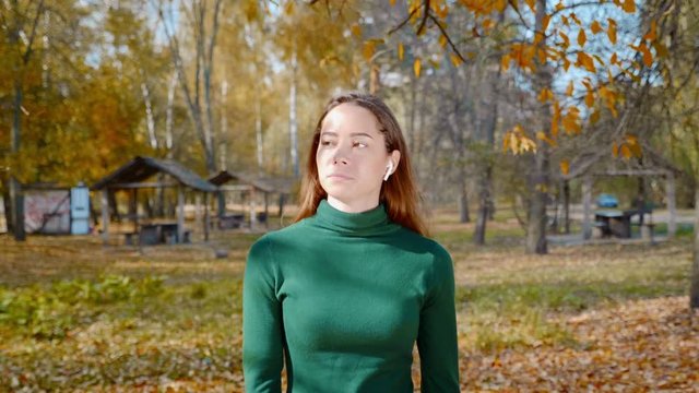 Young attractive brunette woman with wireless earphones is walking in the park on a background of yellowed trees in autumn. Pretty girl in a green sweater strolling in the middle of the forest. 4K