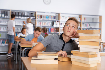 Fototapeta na wymiar Upset schoolboy sitting with stacks of books in library
