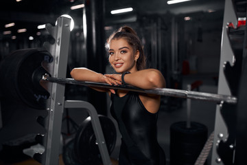 Fototapeta na wymiar Charming smiling fit girl folded hands on metal bar, looking at camera with broad smile, expressing cheerful expression, satisfied by good training in modern fitness studio, portrait, indoor shot