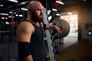 Rude hairless weightlifter with thick beard and serious face, dressed in black t shirt, standing...