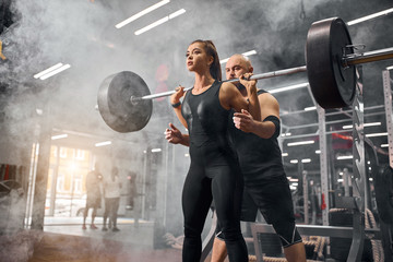 Obraz na płótnie Canvas Low angle shot of sporty friends training in underground garage, standing in white smoke, holding heavy black barbell, brutal professional sportsman supporting young female partner, indoor shot