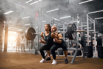 Fototapeta na wymiar Brutal male weightlifter standing behind young sporty woman backing up squatting process with heavy barbell in brightly lighted gym, practicing in white smoke, shot from below, indoor shot