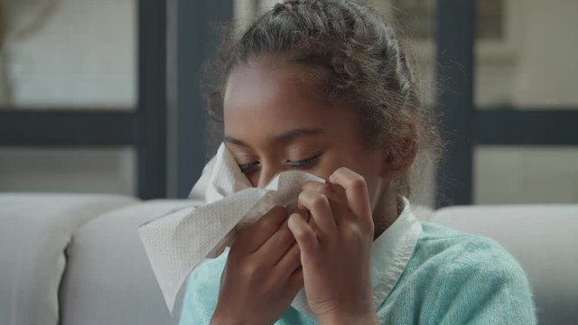 Portrait of adorable elementary age sick african american girl blowing her runny nose into tissue paper at home. Unhealthy child suffering from running nose and sneezing, wiping nose by tissue paper.