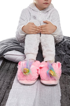 Cropped medium shot of a child in white textured pyjamas and plush house slippers made in the form of pink unicorn with rainbow mane. The child is sitting on the striped carpet and a gray plaid.