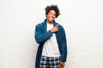 young black man wearing pajamas with gown smiling cheerfully, feeling happy and pointing to the...