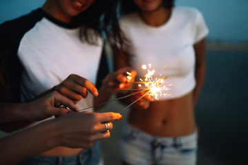 Young happy girls stands on beach with sparkler in sunset light. Teenage girls with sparklers...