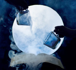 Two hands pour blue smoke from cups onto a book against the backdrop of the moon