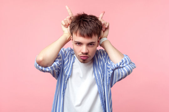 Portrait Of Bully Young Brown-haired Man With Small Beard And Mustache In Casual Striped Shirt Showing Bull Horn Gesture With Fingers, Attack Or Defense. Indoor Studio Shot Isolated On Pink Background