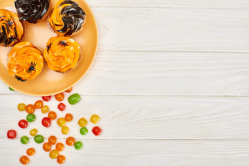 top view of colorful bonbons near orange plate with cupcakes on white wooden table, Halloween treat