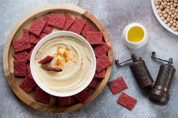 Round wooden serving tray with a bowl of hummus and beetroot crackers, top view over beige stone surface