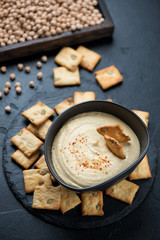 Bowl of hummus and crackers with pumpkin seeds over black stone background, elevated view, vertical shot