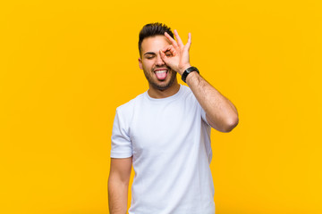 young hispanic man smiling happily with funny face, joking and looking through peephole, spying on...
