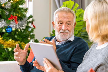 Senior couple using tablet to video phone call to greeting their family for Christmas festival, sitting on sofa with decoration and tree