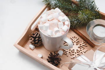 Obraz na płótnie Canvas cocoa with marshmallows, fir branch, cones, candle, snowflakes, gift boxes with white ribbons on a tray on a light