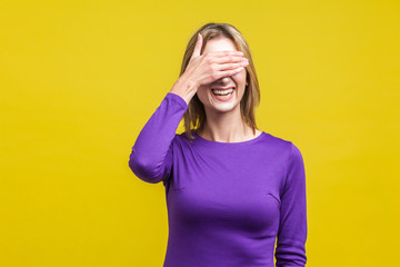 So shameful, I'd rather not watch this! Portrait of bashful positive woman in elegant purple dress covering eyes with hand and laughing, shy to look. indoor studio shot isolated on yellow background