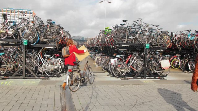 AMSTERDAM, Netherlands - October 2019: young woman on a bicycle rides up to a large bicycle park. A black man walks past.