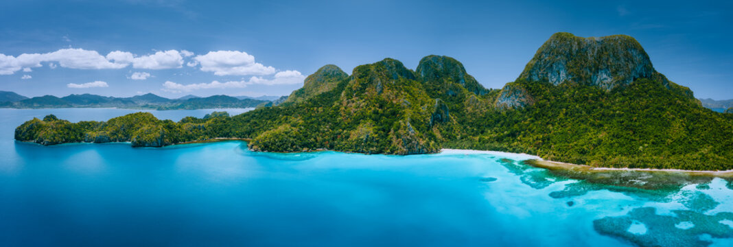 Aerial drone panoramic view of uninhabited tropical island with rugged mountains, rainforest jungle, sandy beaches surrounded by blue ocean