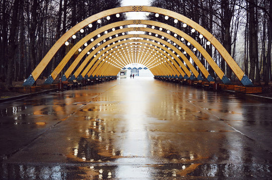 Moscow, Russia. Sokolniki Park In Rainy Autumn Day. Illuminated Wooden Arch With Reflections In Puddles. Two People Walking Far Away