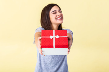 Happy brunette woman in casual holding gift box and looking at the camera while enjoys over yellow background. Young woman hold gift in christmas style