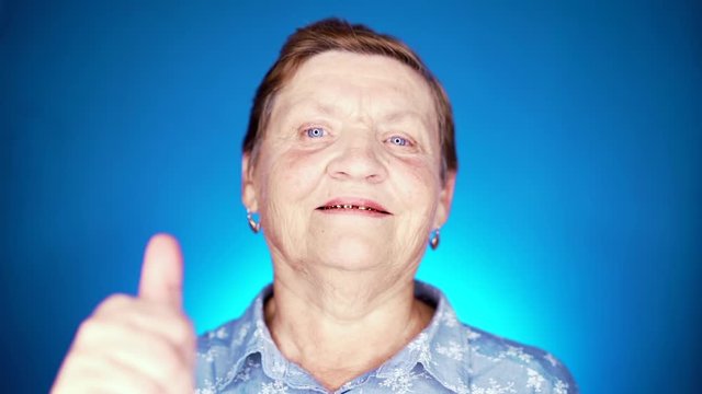 Beautiful portrait of smiling aged woman on blue background. Caucasian grandmother looking at camera and showing thumb-up sign - gesture of approval.