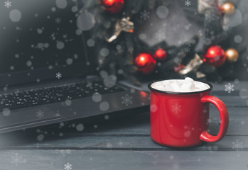 hot chocolate or coffee and marshmallows next to a gray laptop, spruce Christmas wreath with red balls on a gray background with snowflakes and white flakes of snow
