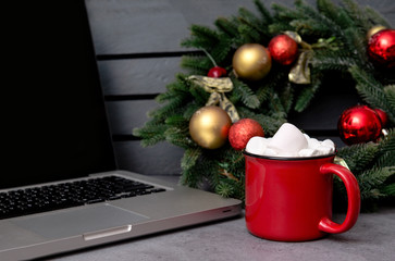 cocoa, hot chocolate and marshmallows next to the laptop, spruce Christmas wreath with  balls on a gray background, side view