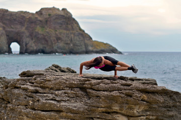 A young athletic woman is doing fitness on a rocky seashore next to a grotto
