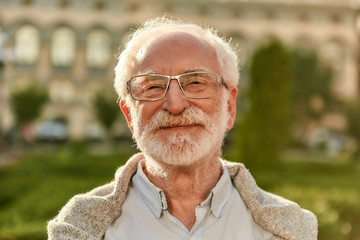 Positive attitude. Portrait of handsome bearded senior man in glasses looking at camera and smiling while spending time outdoors on a sunny day - Powered by Adobe