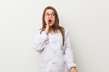 Young doctor woman against a white wall is saying a secret hot braking news and looking aside