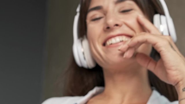 Cropped view of happy cute young woman smiling and touching her chin with hand while listening music with wireless headphones sitting indoors at home