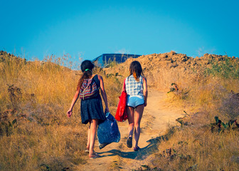 Obraz na płótnie Canvas Two girls with heavy bags climb to the top of the hill. People walking from the beach to the car park on a hot summer day on the sea coast. Overcoming difficulties concept.