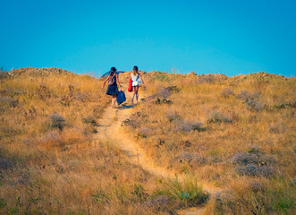 Two girls with heavy bags climb to the top of the hill. People walking from the beach to the car park on a hot summer day on the sea coast. Overcoming difficulties concept.