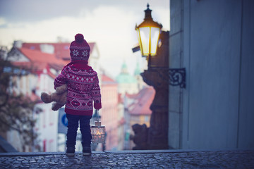 Beautiful toddler child with lantern and teddy bear, casually dressed, looking at night view of Prague city